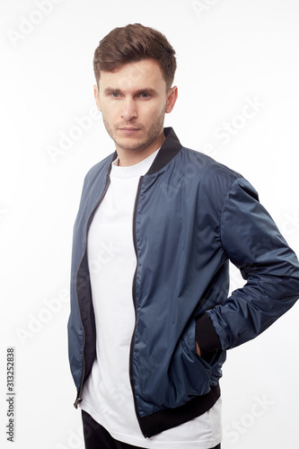 Leinwand Poster Young european man in white sweater and black pants, blue bomber jacket posing on white background