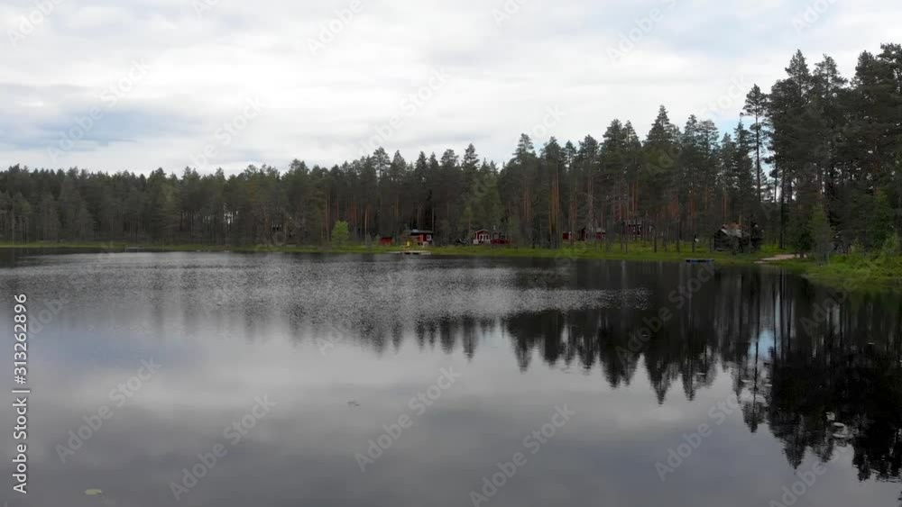 Drone flight through pine tree forest towards a lake with some wooden cabins located at the edge in Dalarna county, Vansbro municipality in Sweden