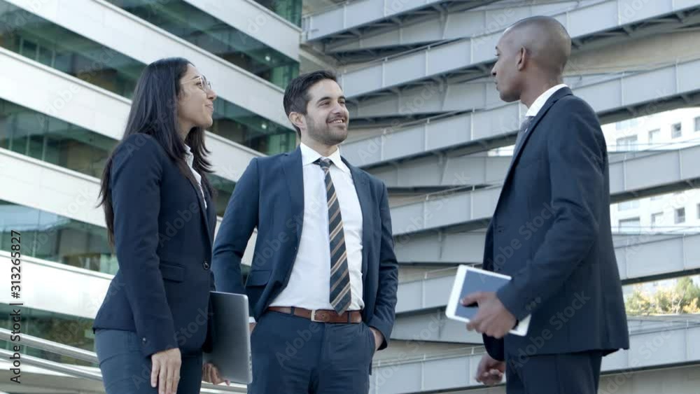 Coworkers with digital devices greeting each other. Low angle view of young business colleagues holding laptop with digital tablet and shaking hands on street. Business concept Coworkers with digital devices greeting each other. Low angle view of young business colleagues holding laptop with digital tablet and shaking hands on street. Business concept
