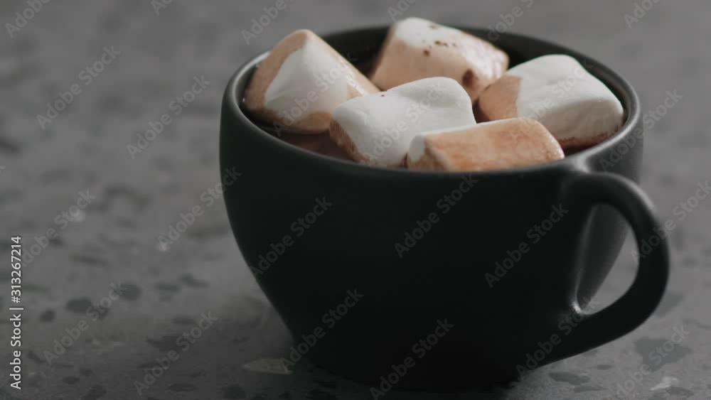 Slow motion sprinkle cocoa powder on marshmallow cocoa drink in a black cup on terrazzo countertop Slow motion sprinkle cocoa powder on marshmallow cocoa drink in a black cup on terrazzo countertop