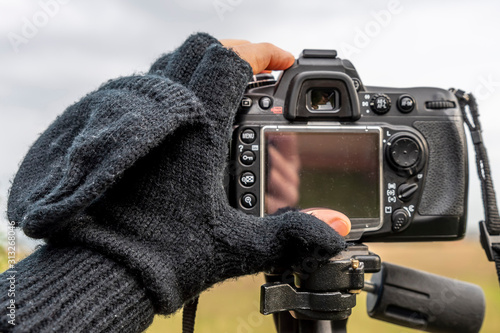 A hand covered in a woolen glove with only the fingertips uncovered adjusts a camera on a tripod in the winter season