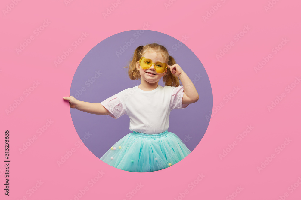 Surprised Little Child girl in heart shap sunglasses looking out of a pink round hole. Banner. Happy girl Looking at camera. Wow funny face Surprised Little Child girl in heart shap sunglasses looking out of a pink round hole. Banner. Happy girl Looking at camera. Wow funny face