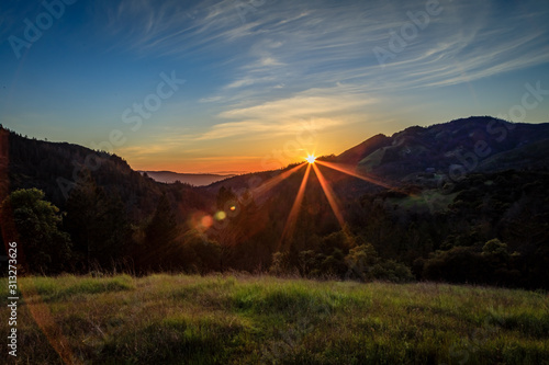 The edge of the sun creates a lens flare as it slips behind the edge of a mountain with wispy blue skies in the background at Sugarloaf Ridge State Park