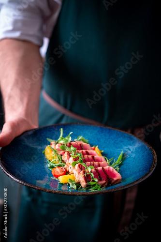 Man holds a plate of Sliced grilled tuna with green salad and tomatoes.