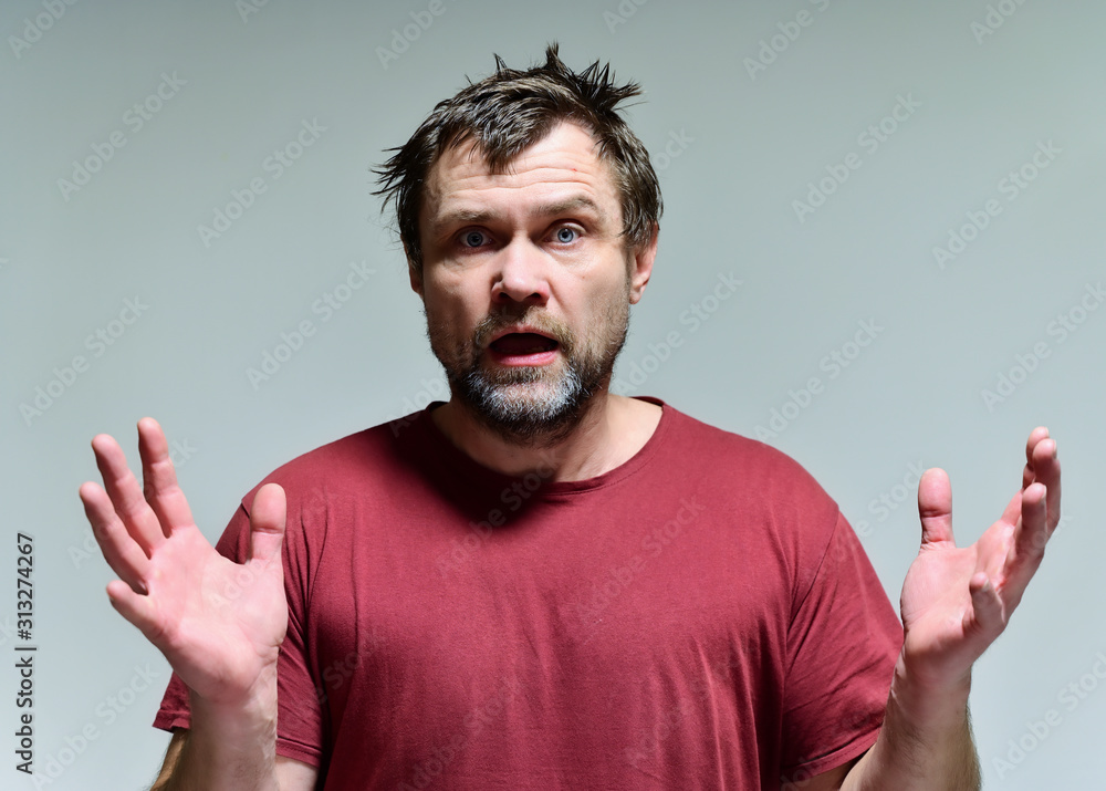Portrait of a wild unkempt unshaven middle-aged man of 40 years in a burgundy t-shirt on a gray background. He stands right in front of the camera, talking, showing emotions. Waves his hands. Portrait of a wild unkempt unshaven middle-aged man of 40 years in a burgundy t-shirt on a gray background. He stands right in front of the camera, talking, showing emotions. Waves his hands.