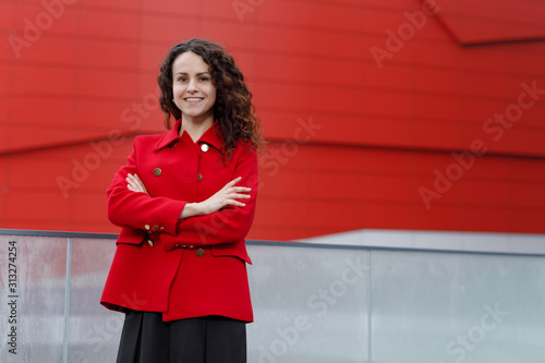 Cheerful young woman with curly hair, with a beautiful smile, keeps her hands crossed, dressed in a red jacket. Picture of a business woman, looks confidently at the camera. photo