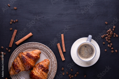 Croissants on a ceramic plate, a cup of coffee, cinnamon sticks and coffee beans on a black wooden table. Top view with space for text. Flat lay composition. Background for restaurant, bakery, cafe.