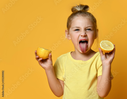 Little smiling blond girl in yellow t-shirt holding halves of fresh lemon, showing tongue and feeling sour over yellow background