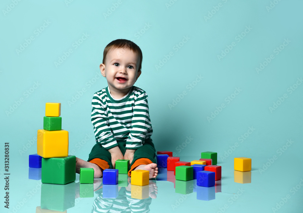 Little baby boy in stylish casual clothing barefoot sitting on floor, playing with colorful toy cubes, showing tongue Little baby boy in stylish casual clothing barefoot sitting on floor, playing with colorful toy cubes, showing tongue