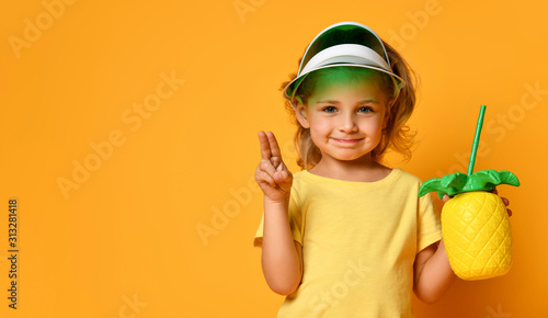 Little smiling cute blond girl in yellow t-shirt and hat holding fresh healthy fruit juice in pineapple shaped bottle with straw