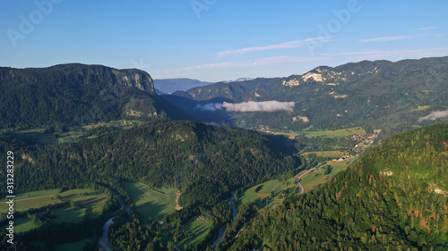 Aerial view of Bled Lake valley. Natural landscape with green meadows, fields, hills and mountains, small houses and trees. Sunny blue sky. Sun rays.