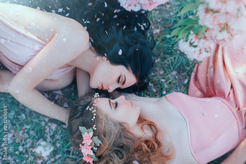 portrait two young women lie cuddling on grass in spring garden, luxurious long curly hair strewn with flowers rose petals. Attractive face gentle makeup. Different sisters. Natural cosmetics concept - obrazy, fototapety, plakaty 