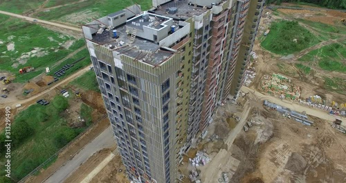 An aerial view of an almost finished residential multi storey building standing on the construction site. There are construction materials and some bulders on the dug ground. The background is filled photo