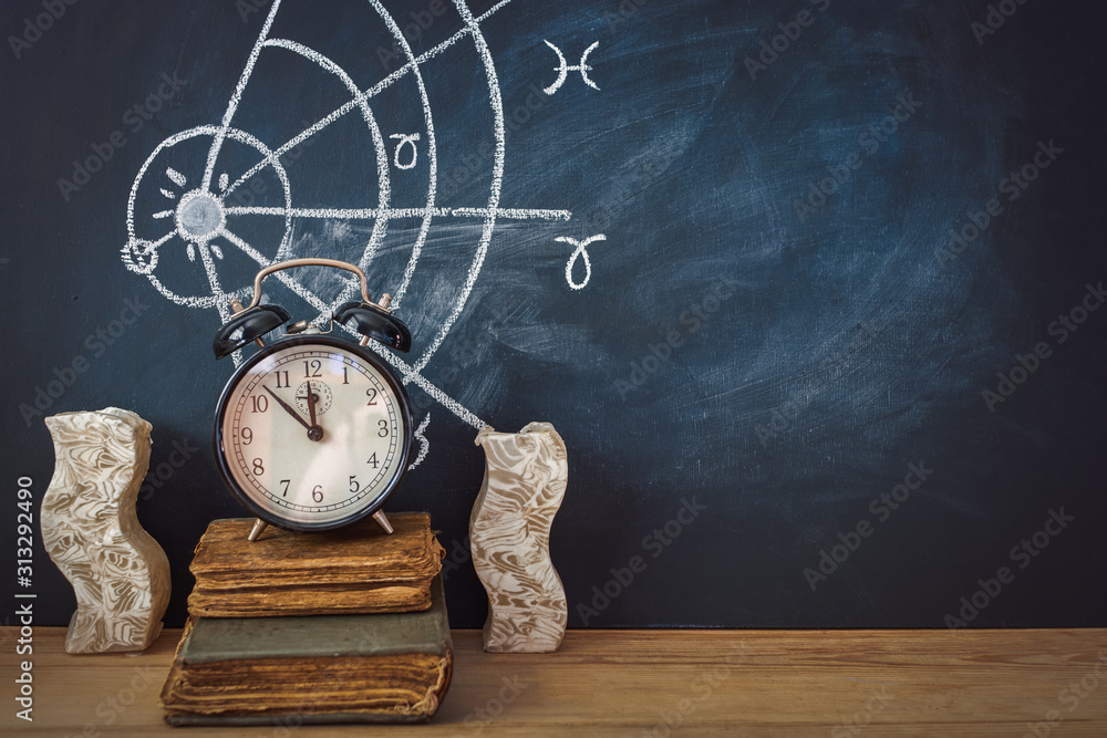 Alarm clock and old books next to a candle on a horoscope background drawn on a chalkboard