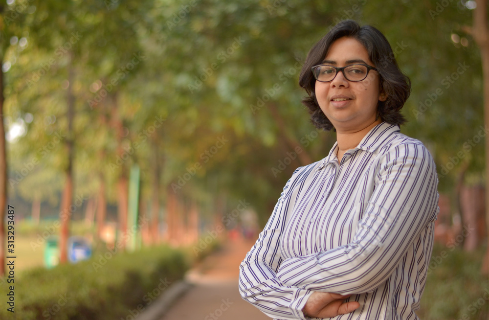 Closeup portrait of a confident young Indian Corporate professional woman with short hair and spectacles, crossed folded hands in an outdoor setting wearing a black white stripped shirt in a park Closeup portrait of a confident young Indian Corporate professional woman with short hair and spectacles, crossed folded hands in an outdoor setting wearing a black white stripped shirt in a park