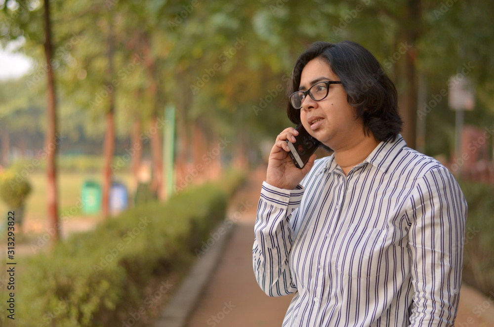 Portrait of a confident looking young Indian woman with short hair and spectacles speaking on a mobile phone and holding a laptop in outdoor setting wearing a formal striped shirt in the park Portrait of a confident looking young Indian woman with short hair and spectacles speaking on a mobile phone and holding a laptop in outdoor setting wearing a formal striped shirt in the park