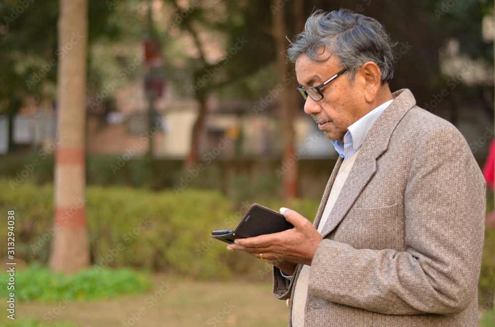 Obraz premium Lifestyle shot of senior Indian man wearing a tweed coat working on a phone in a park in New Delhi, India Concept - Digital India and digitally literate citizens