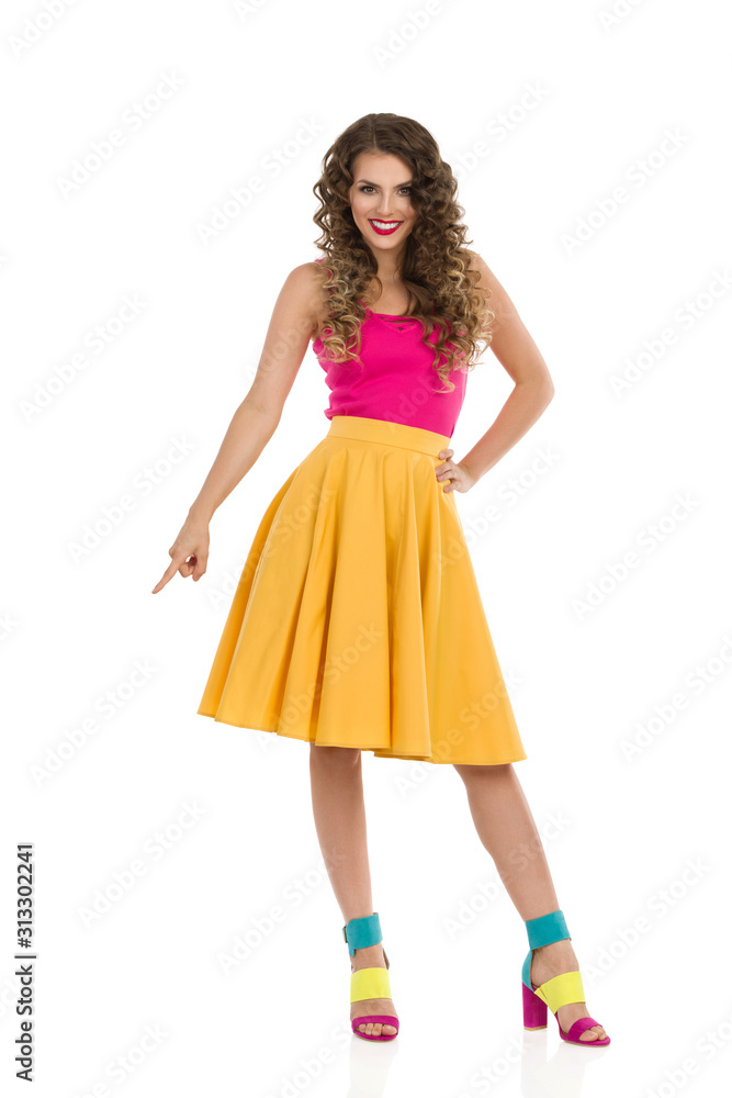 Smiling Young Woman In Colorful High Heels, Vibrant Skirt And Pink Top Is Pointing Down.