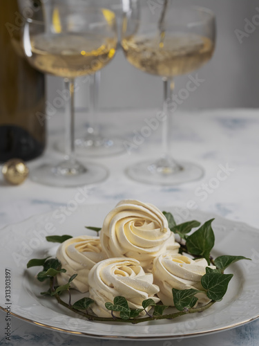 Apricot marshmallow surrounded by an ivy branch is located in a white plate, in the background are glasses of wine. Selective focus on the home zephyr, vertical orientation. Delicious homemade sweets.
