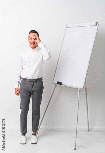 Young latin coaching man with a whiteboard isolated excited keeping ok gesture on eye.