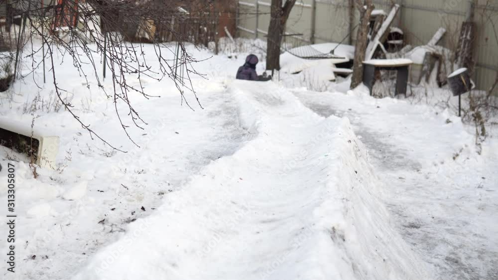 A child rolls on a snow slide. Winter fun on the street. In the cold winter, the child is dressed in a jacket with a scarf. Back view A child rolls on a snow slide. Winter fun on the street. In the cold winter, the child is dressed in a jacket with a scarf. Back view