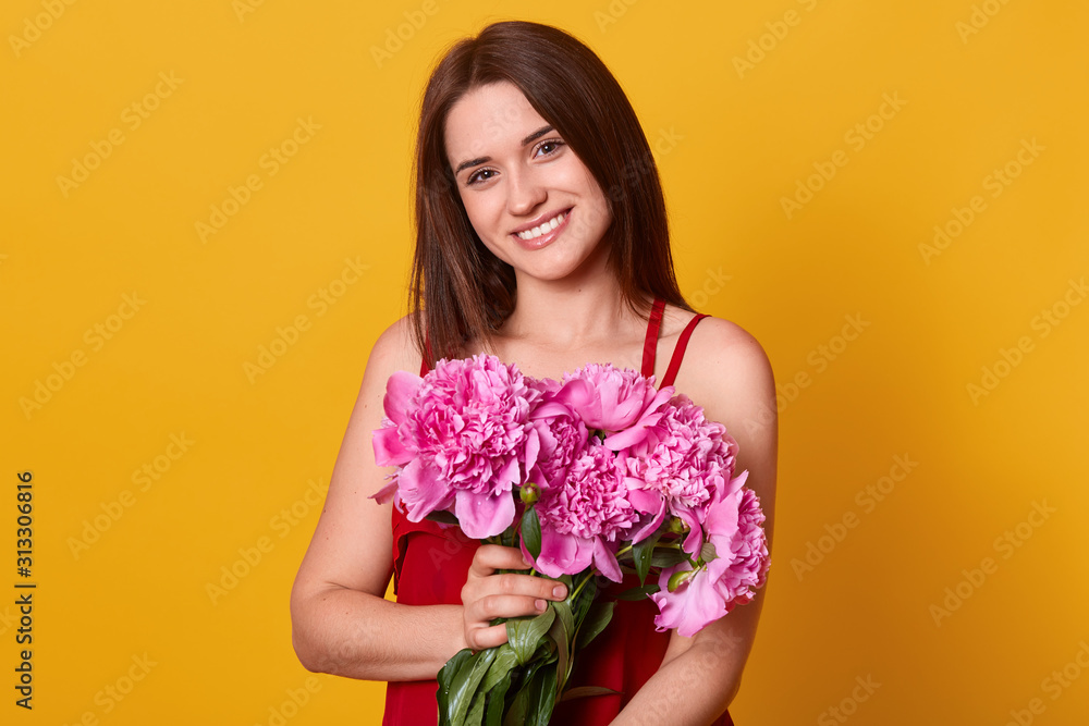 Smiling cute female posing isolated over yellow background in studio, looking directly at camera with charming smile, holding rosy peonies in hands, wearing red dress, having straight dark hair.