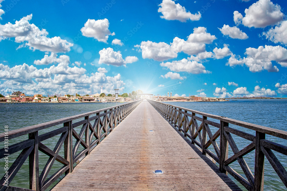 Long wooden bridge leading over a sea lagoon. It leads to Fadiouth Island in Senegal, Africa. Sun rays shine from the end of the bridge. It is a beautiful natural background.