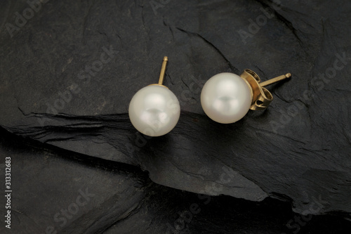 A close up of a pair of white pearl earring studs with yellow gold post and nut on a rough black slate background.