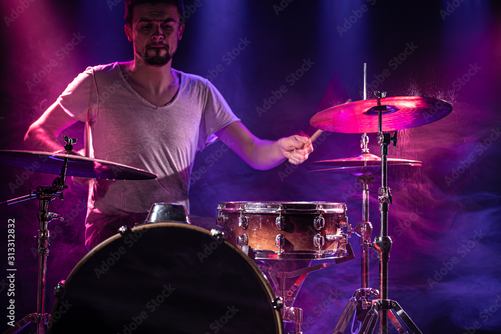 Obraz premium The drummer plays the drums. Beautiful blue and red background, with rays of light. Beautiful special effects smoke and lighting. The process of playing a musical instrument. Close-up photo.