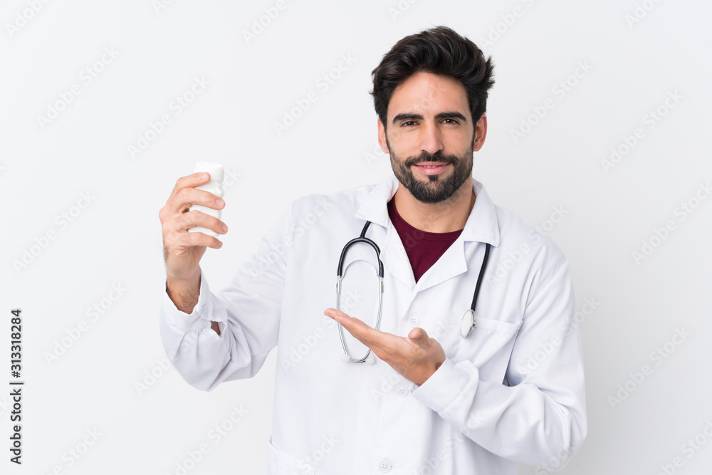 Young handsome man with beard over isolated white background wearing a doctor gown and holding pills Young handsome man with beard over isolated white background wearing a doctor gown and holding pills