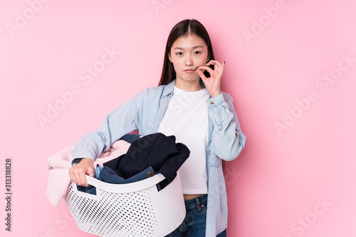 Young chinese woman picking up dirty clothes isolated with fingers on lips keeping a secret. photo