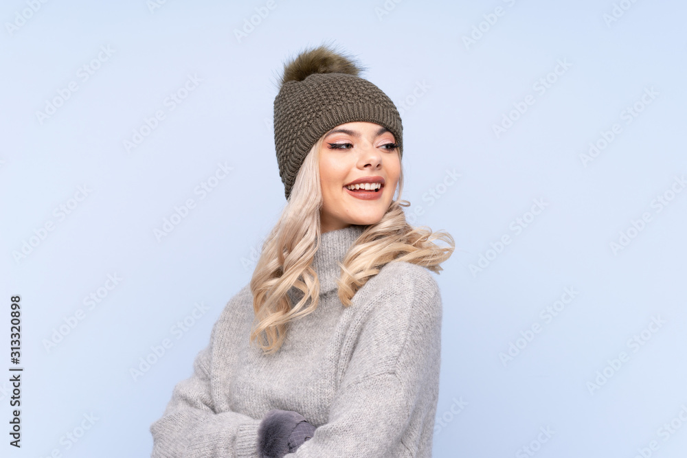 Young teenager girl with winter hat over isolated blue background laughing