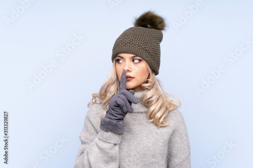 Young teenager girl with winter hat over isolated blue background doing silence gesture