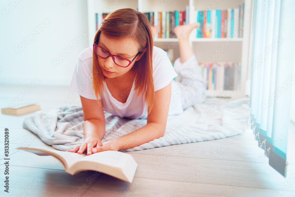 Portrait of young beautiful woman girl student reading or study at home lying on the floor in front of the book shelves wearing glasses holding a book Portrait of young beautiful woman girl student reading or study at home lying on the floor in front of the book shelves wearing glasses holding a book