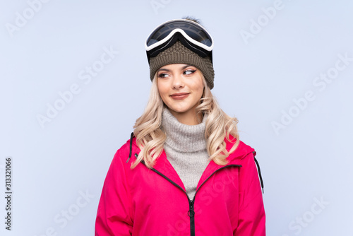 Skier teenager girl with snowboarding glasses over isolated blue background standing and looking to the side © luismolinero