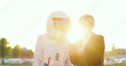 Pretty Caucasian young businesswoman showing something on the screen of smartphone to the man in the astronaut costume while they standing outside in sunlight at the parking. photo