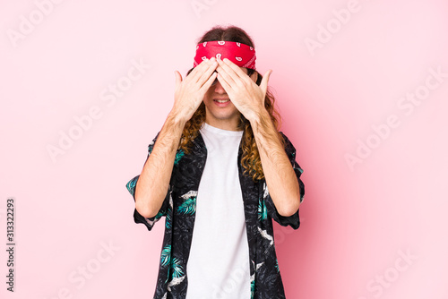 Young long hair hippie man posing in a pink background covers eyes with hands, smiles broadly waiting for a surprise.