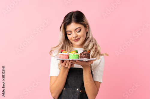 Teenager girl over isolated pink background holding mini cakes enjoying the smell of them