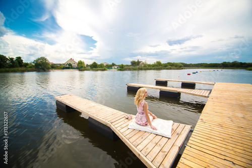 Young charming blond girl sits alone on a wooden pier and looks at the river