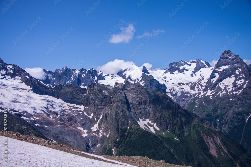 Beautiful panorama of the Caucasus mountains. The top of the mountain range covered with snow. Forest on the slope. Sunny day. Background image for travel and nature.