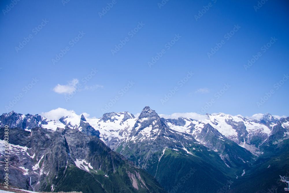 Beautiful panorama of the Caucasus mountains. The top of the mountain range covered with snow. Forest on the slope. Sunny day. Background image for travel and nature.