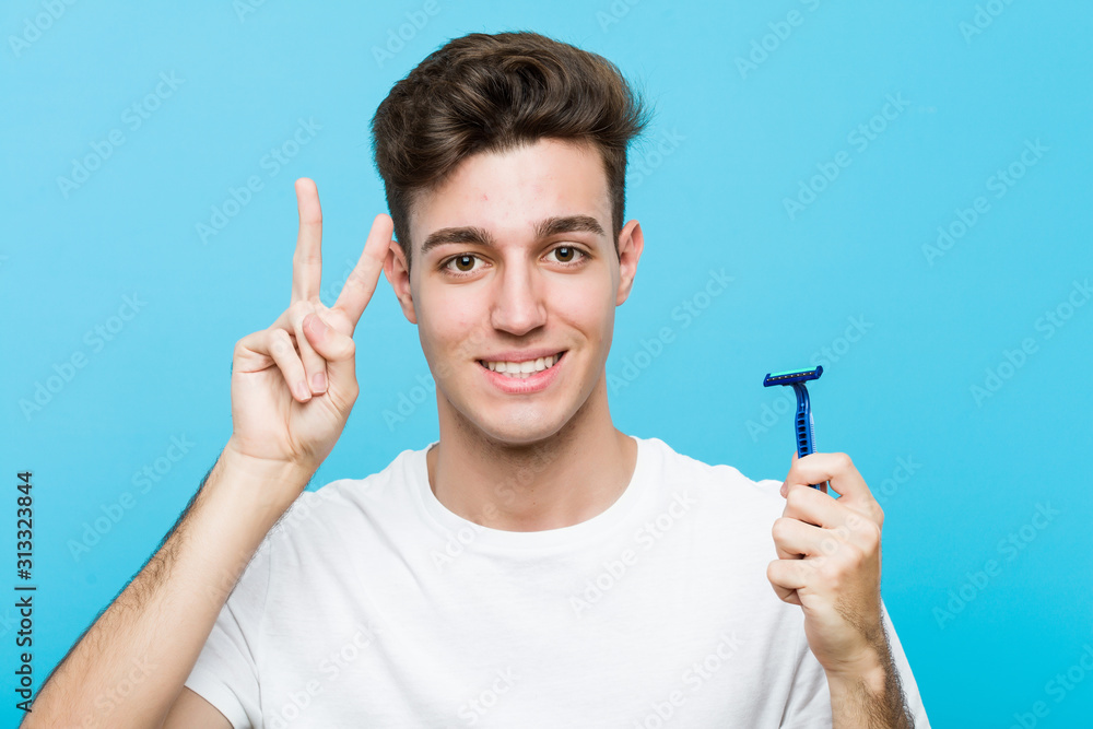 Young caucasian man holding a razor blade showing victory sign and smiling broadly.