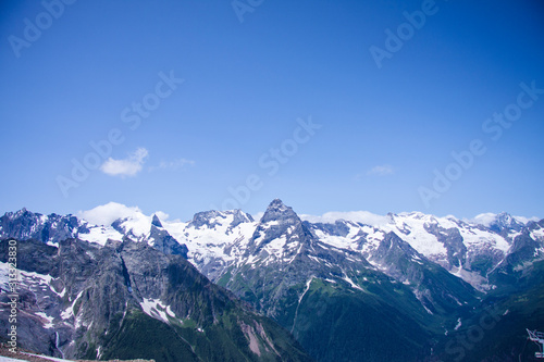 Beautiful panorama of the Caucasus mountains. The top of the mountain range covered with snow. Forest on the slope. Sunny day. Background image for travel and nature.