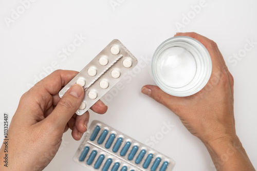 Woman taking pills, female hands holding a blister of white pills, capsules and a glass of water on white table, healthcare and flu treatment concept