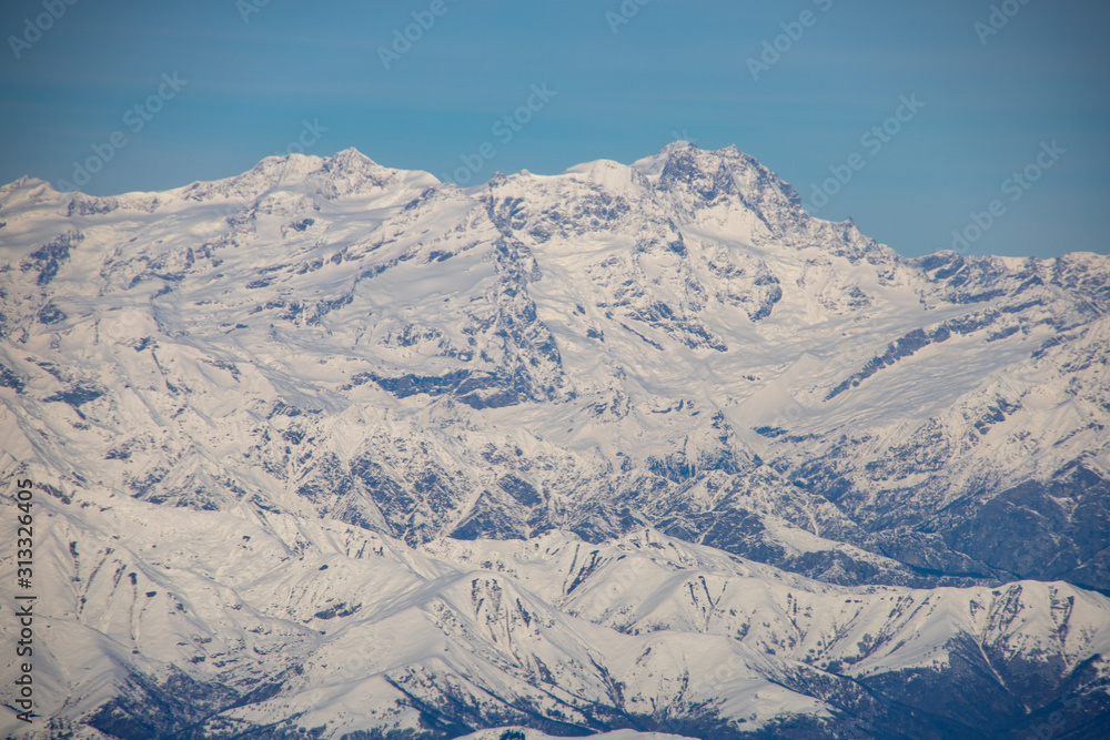 Airplane window photography: overflying the alps, snowy mountains view from above. Blue sky.