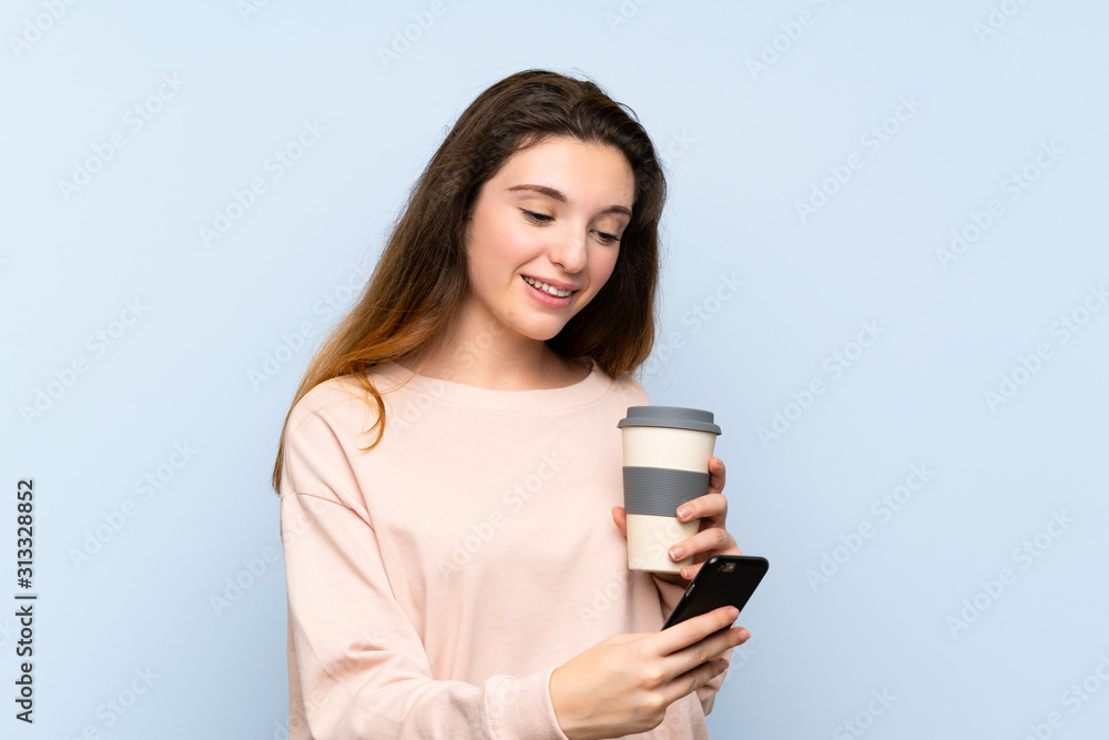 Young brunette girl over isolated blue background holding coffee to take away and a mobile Young brunette girl over isolated blue background holding coffee to take away and a mobile