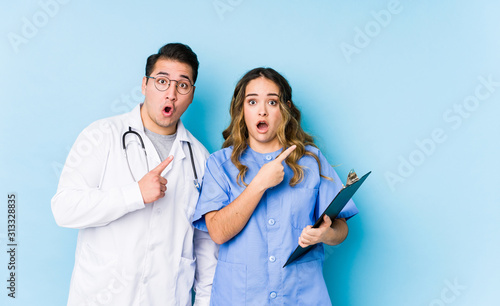 Young doctor couple posing in a blue background isolated pointing to the side