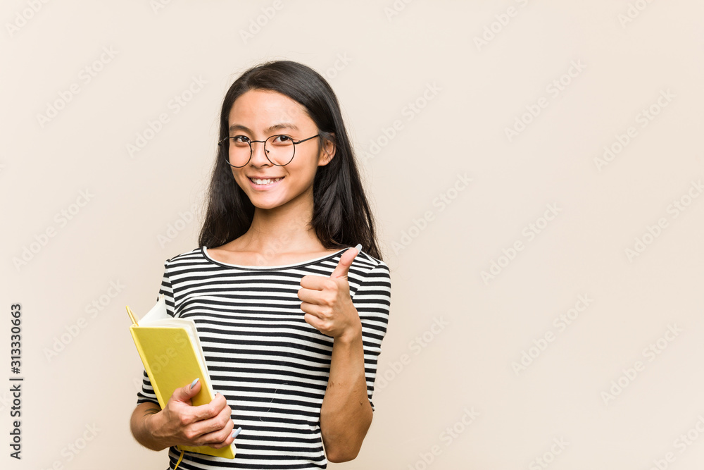 Young asian woman student holding a book smiling and raising thumb up Young asian woman student holding a book smiling and raising thumb up