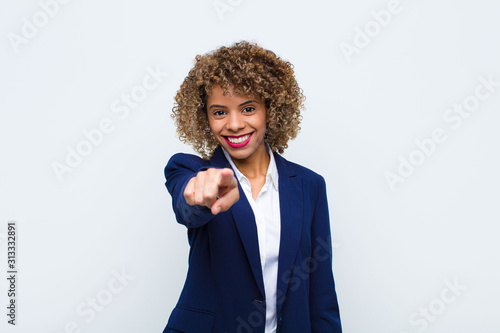 young woman african american pointing at camera with a satisfied, confident, friendly smile, choosing you against flat wall