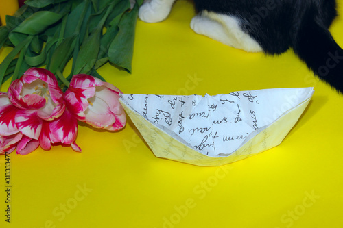White paper boat, pink tulips, blurred silhoette of a cat, yellow background. Abstract colorful background. 